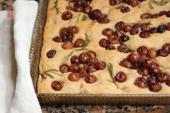 Partial view of focaccia with roasted grapes on metal pan, kitchen towel beside it
