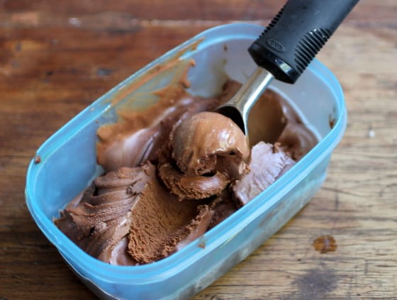 Blue plastic container with chocolate ice cream and scoop on wooden table.