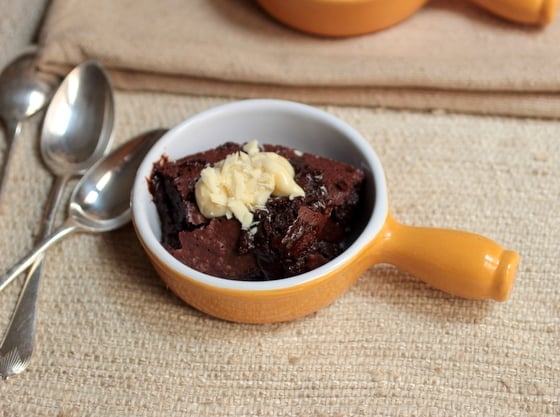 Top view of yellow shallow ramekin with chocolate pudding on a sand colored cloth, spoons on the side