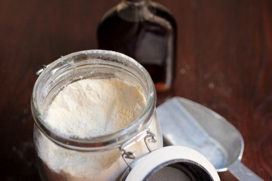 Pancake mix in a mason jar, a metal scoop, syrup jar on a wooden table. 