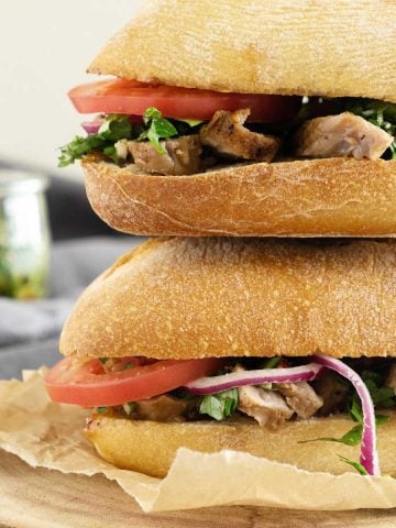 Two steak, tomato and chimichurri sandwiches stacked on a wood board.