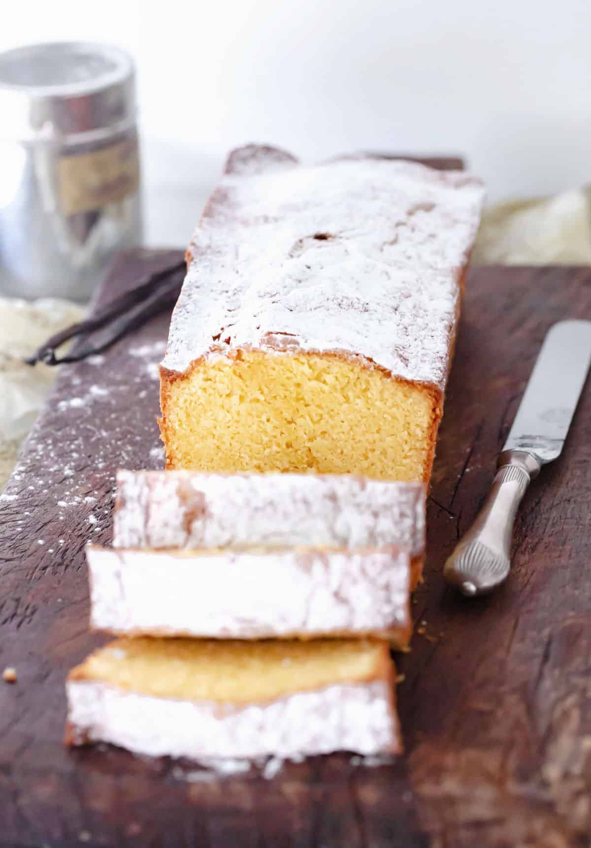 Whole powdered sugar dusted pound cake with cut slices on a dark wooden board. A knife and sugar shaker. 