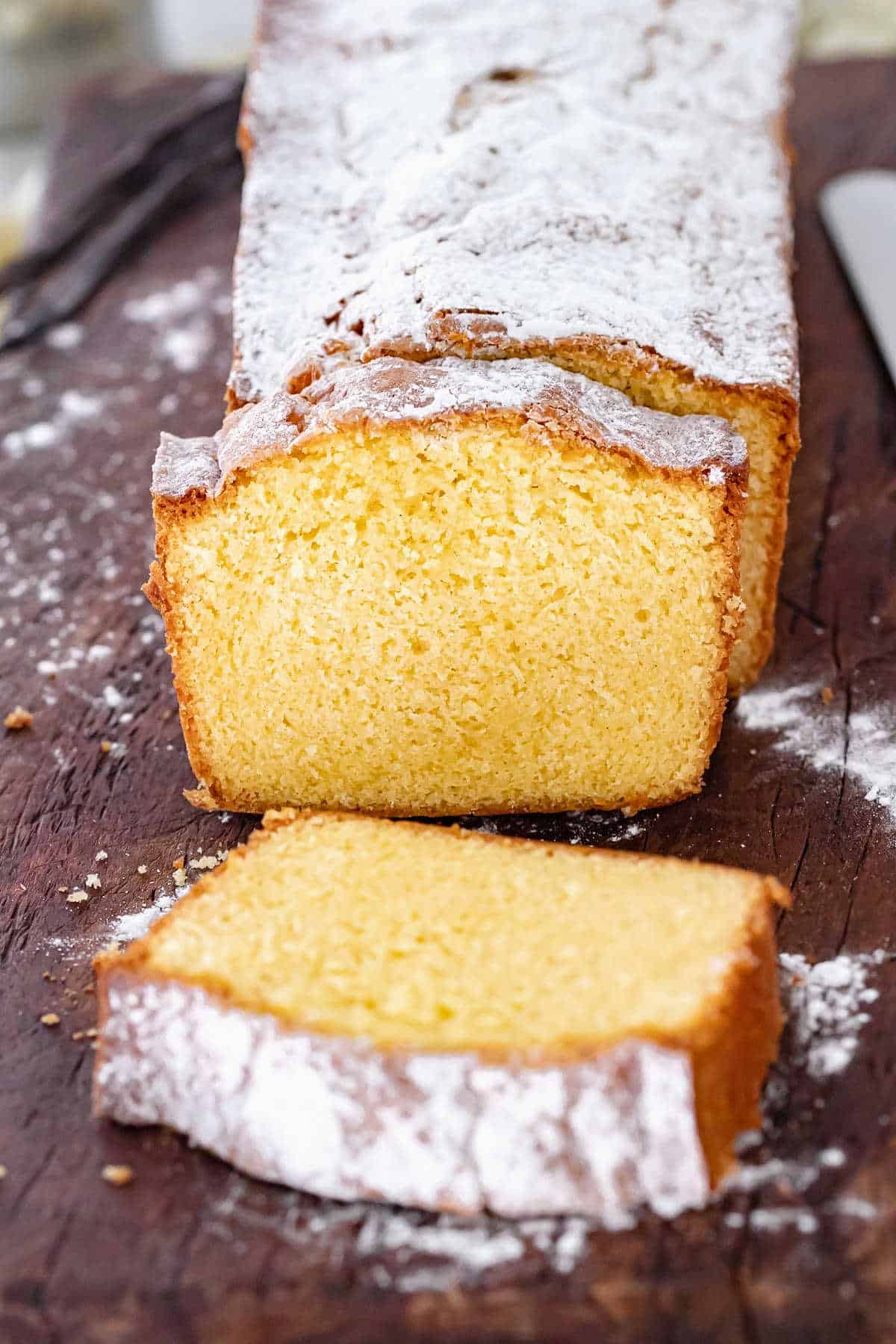 Loaf butter cake with powdered sugar on dark wooden board. Two cut slices.