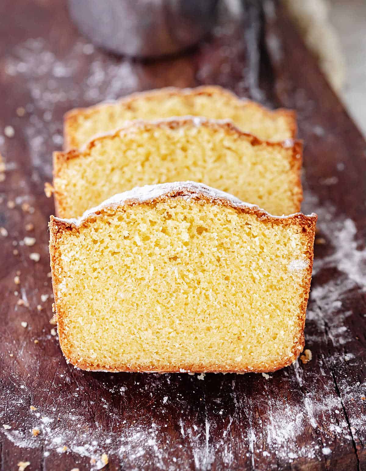 Three slices of vanilla pound cake on a dark wooden board with powdered sugar spots.