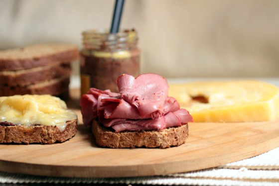 Open pastrami and cheese sandwich on a wooden board. Stack of bread slices and mustard jar in the background. 