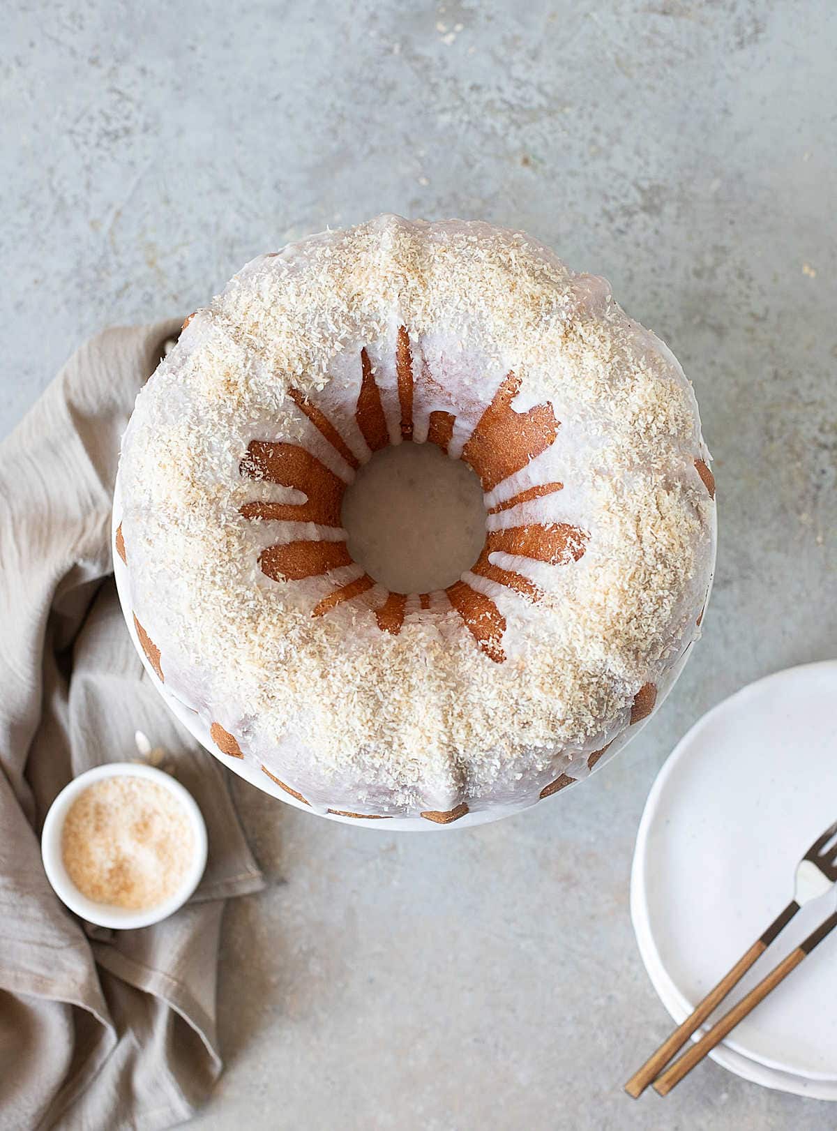 Top view of glazed coconut mango bundt cake. Grey surface, cloth, stack of plates.