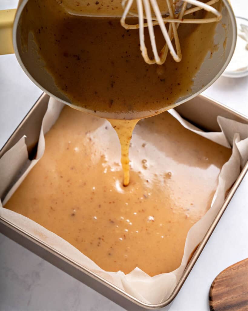 Pouring caramel layer on a square pan with parchment paper. Light gray background.