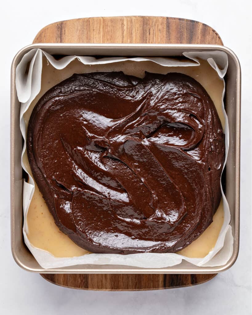 Spreading chocolate ganache on a square pan with caramel and parchment paper. Wooden board, light gray surface.