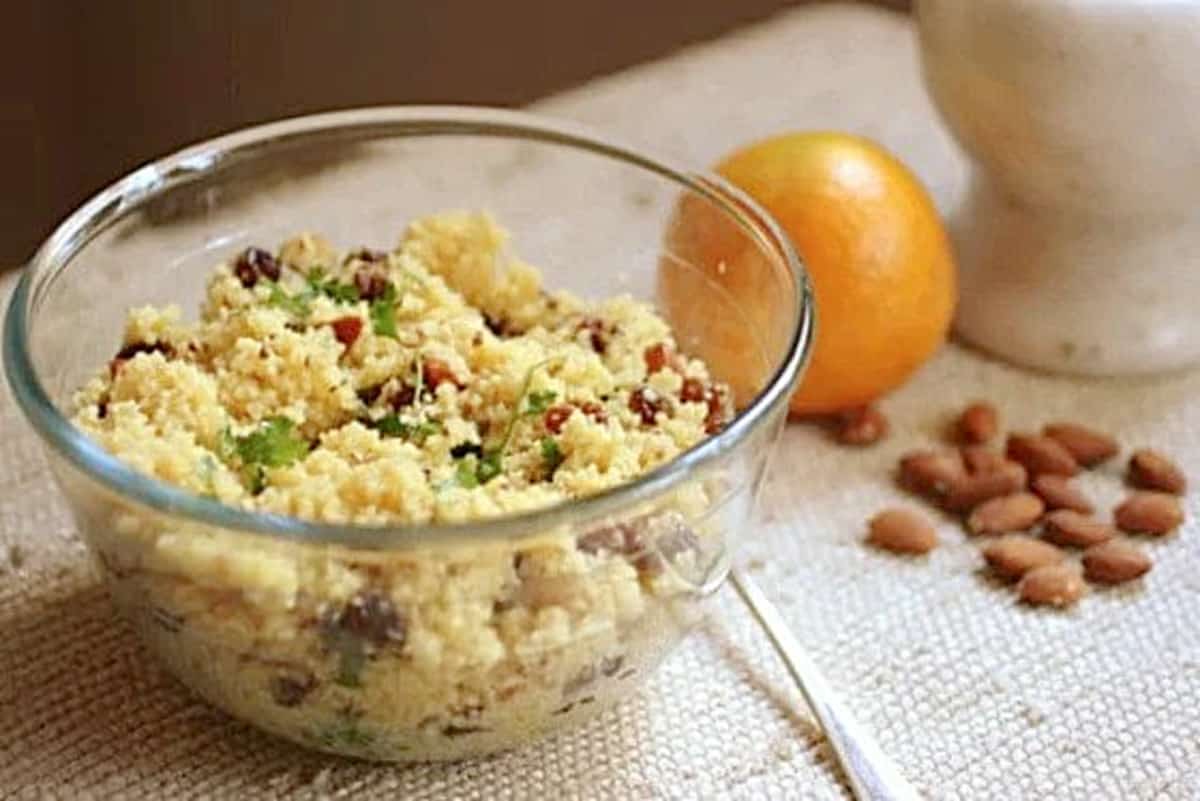 Glass bowl with raisin couscous on beige cloth. Orange and almonds around. 