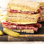 Halved pastrami gruyere rye sandwich on a wooden board. Whole gherkin.