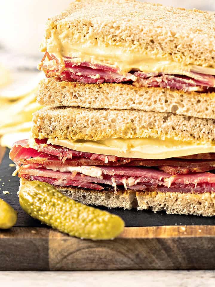 Halved pastrami gruyere rye sandwich on a wooden board. Whole gherkin.