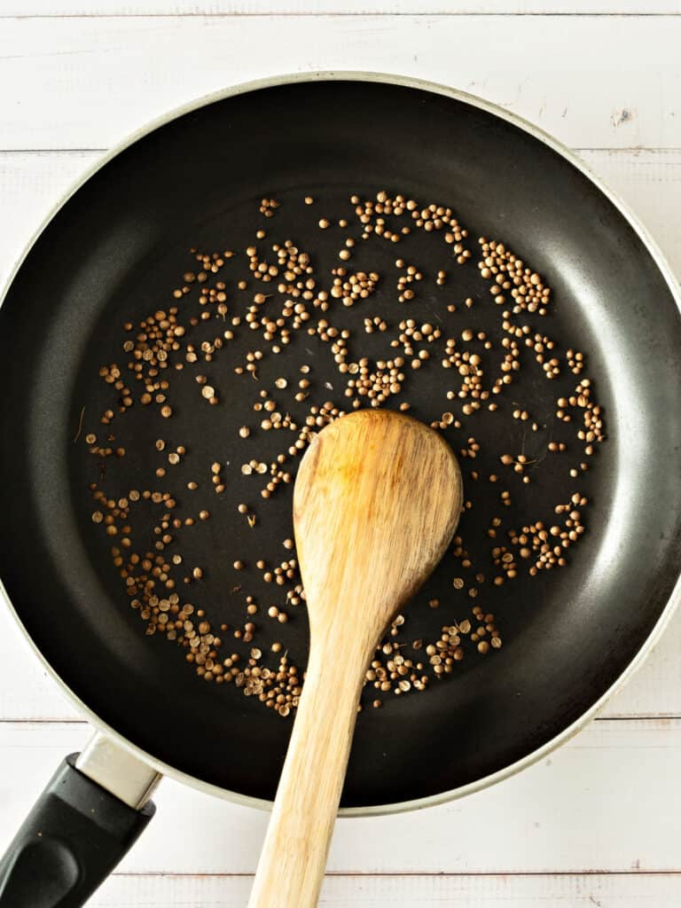 Coriander seeds in a black skillet. Wooden spoon. White surface.