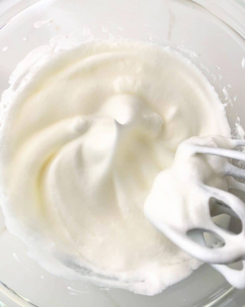 Egg whites beaten to stiff peaks with an electric mixer in a glass bowl. White surface.