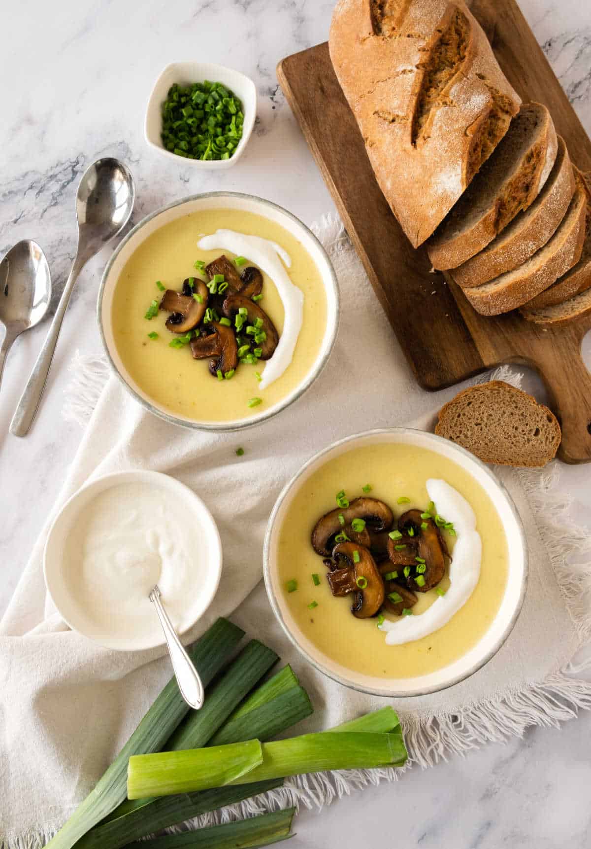 White potato soup bowls with mushrooms, bread loaf on wooden board, white marble with beige linen. Bowl of cream, leeks.
