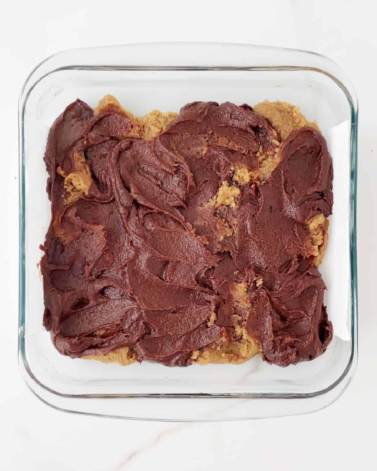 Marbled cookie and brownie dough in a glass square dish on a white marble surface.
