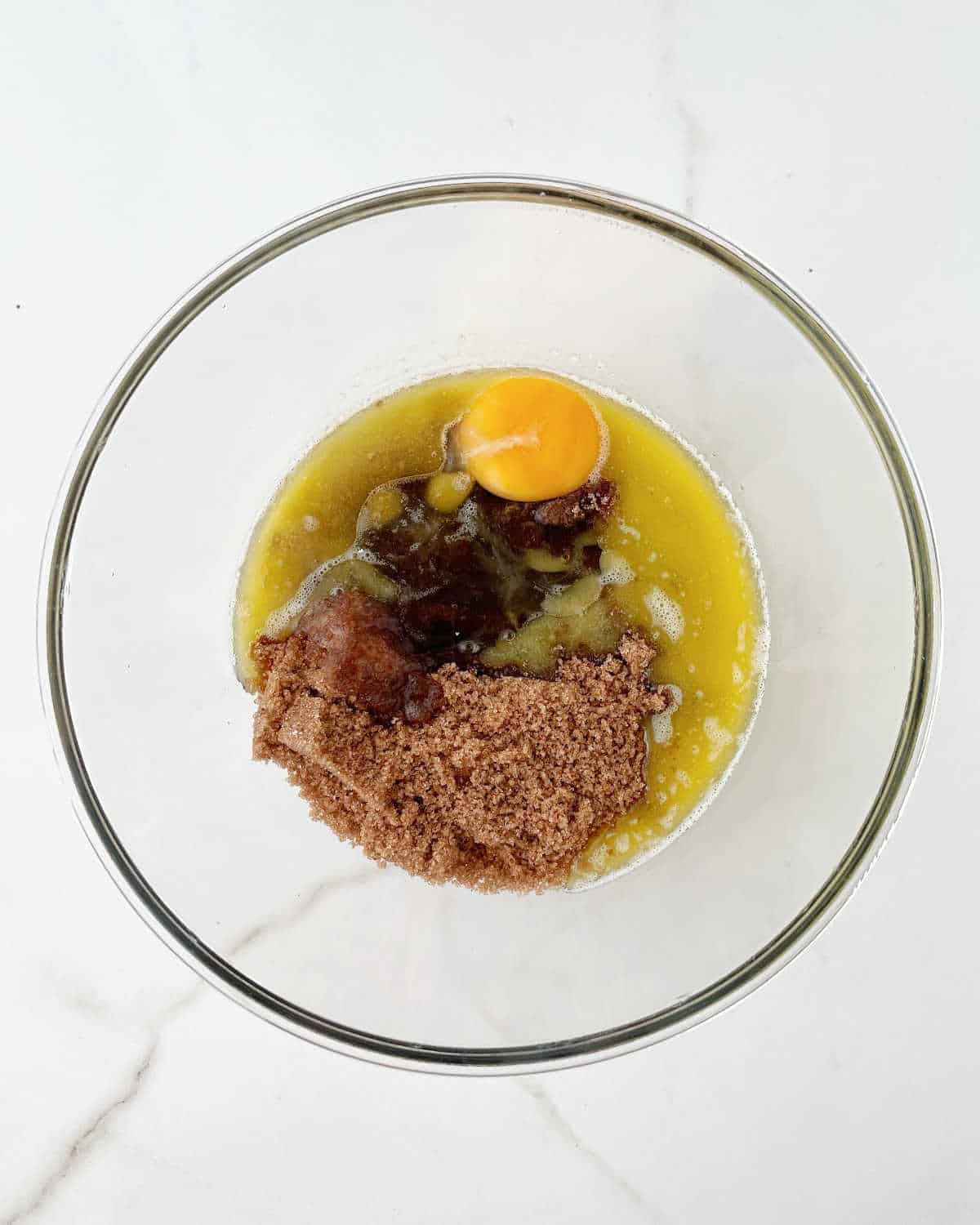 Glass bowl with brown sugar, egg, and melted butter on a white marble surface. 