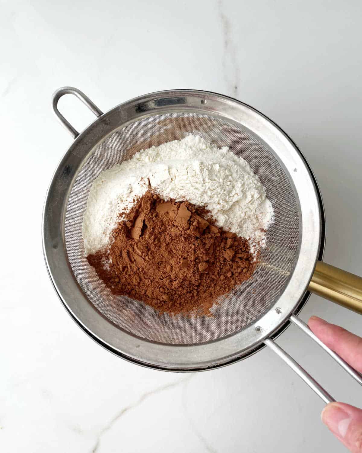 Sifting flour and cocoa powder over a glass bowl. White marble surface.