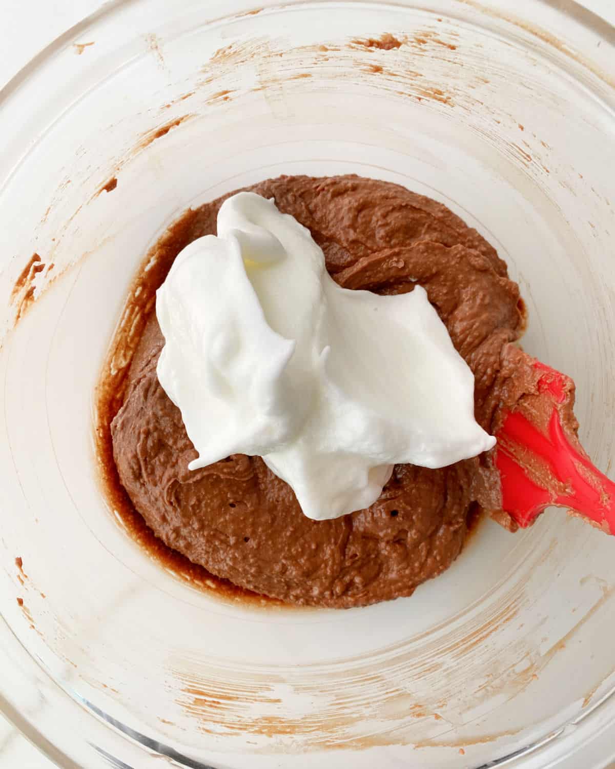 Mound of beaten egg whites added to chocolate cake batter in a glass bowl with a red spatula.