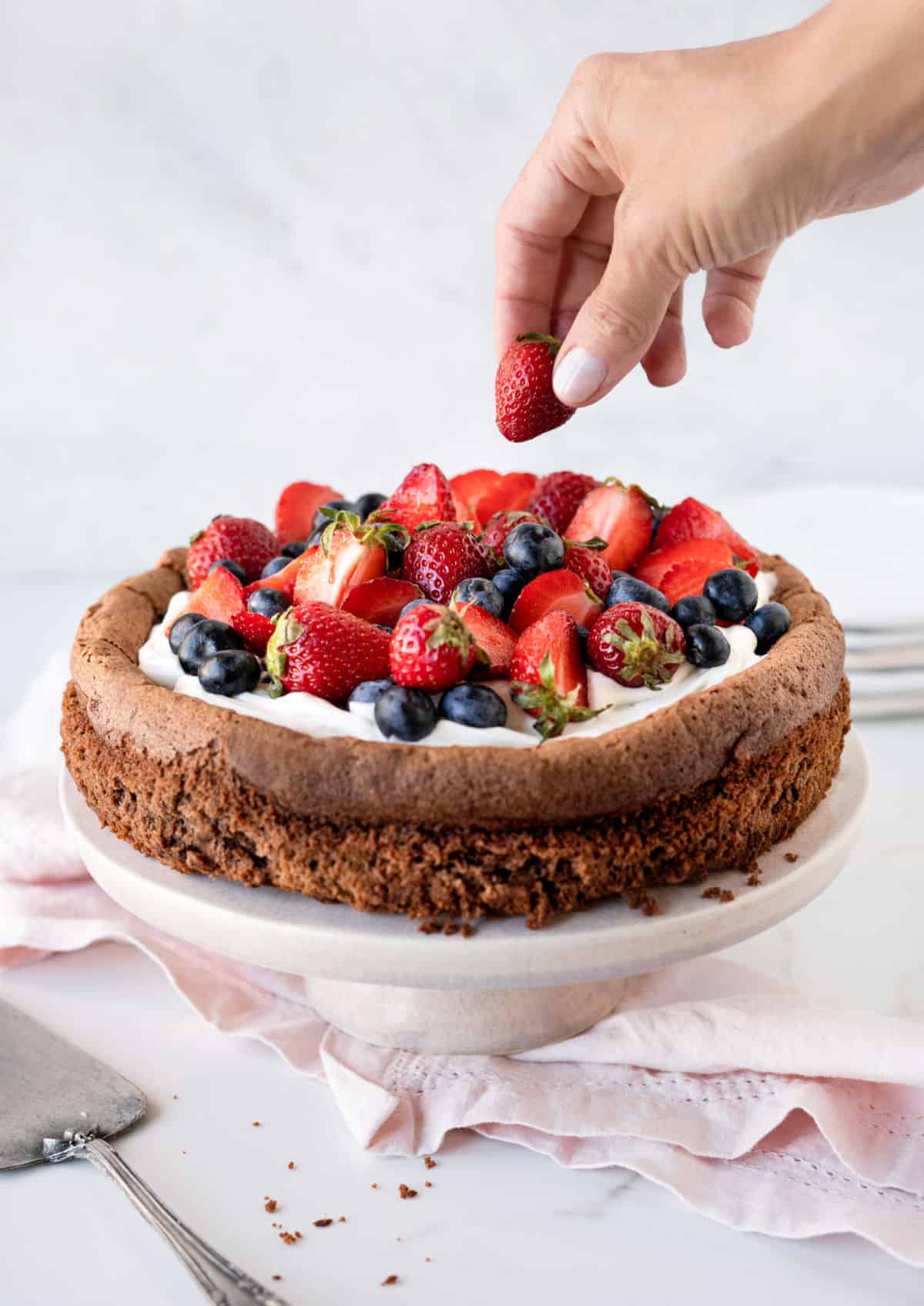 Hand placing strawberry on top of berry topped chocolate cake on a pink cake stand. Pink and white cloth and background. 