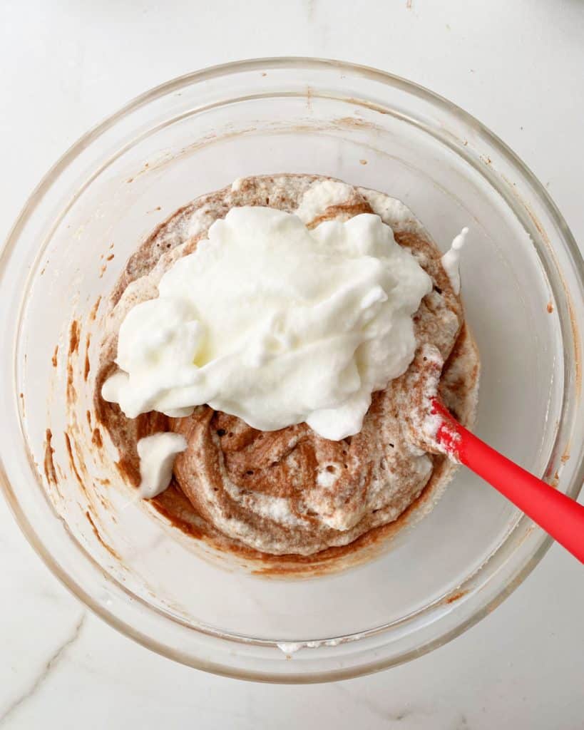 Glass bowl with egg whites added to chocolate cake batter. A red spatula. White marble surface.