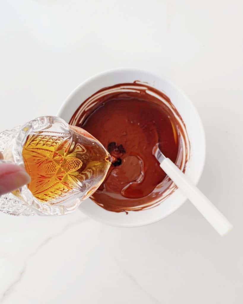 Pouring coffee liquor into white bowl with melted chocolate. White marble surface.