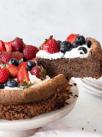 A berry topped chocolate torte on a pink cake stand with one slice being removed. Grey and white background.