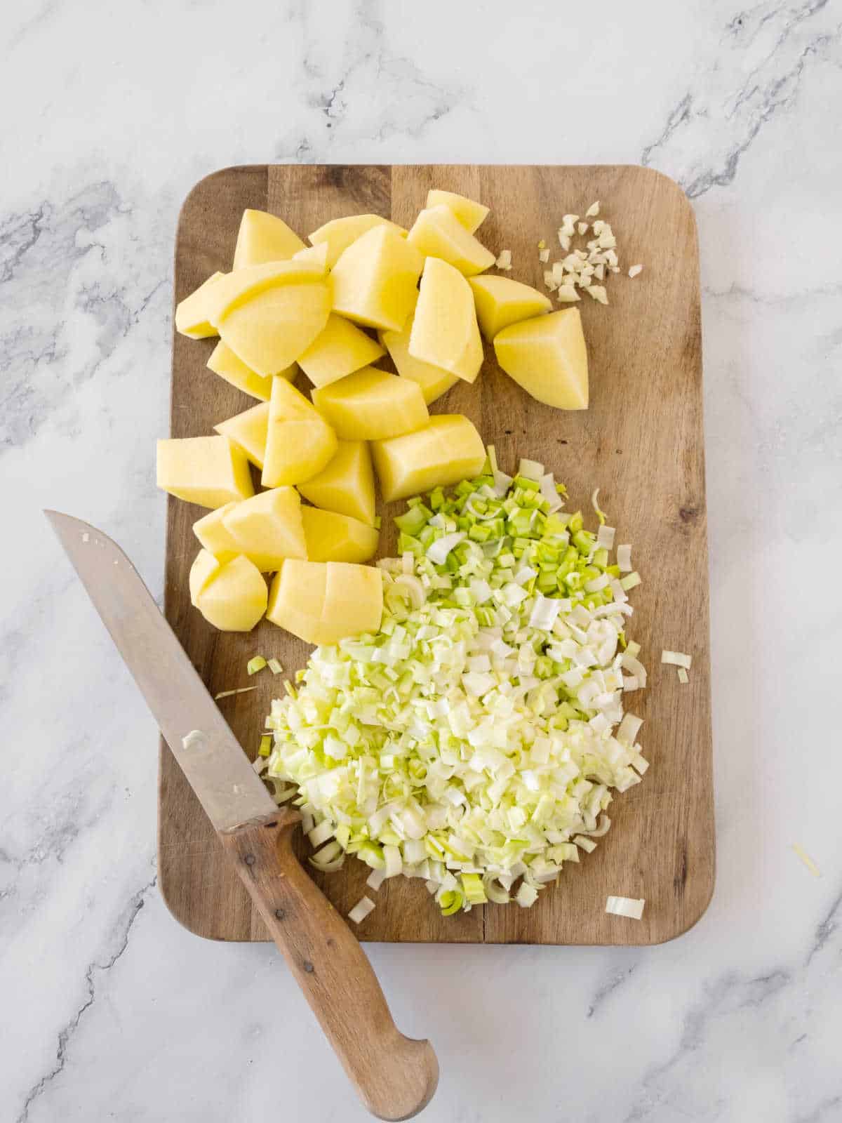 Chopped leeks, garlic and potato pieces on a wooden board. White marbled surface.