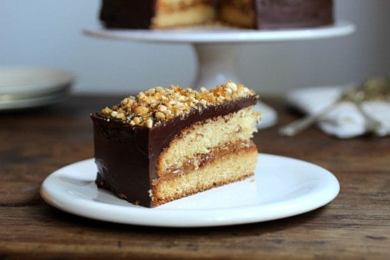 Slice of dulce de leche cake with chocolate frosting on a white plate. Wooden surface.