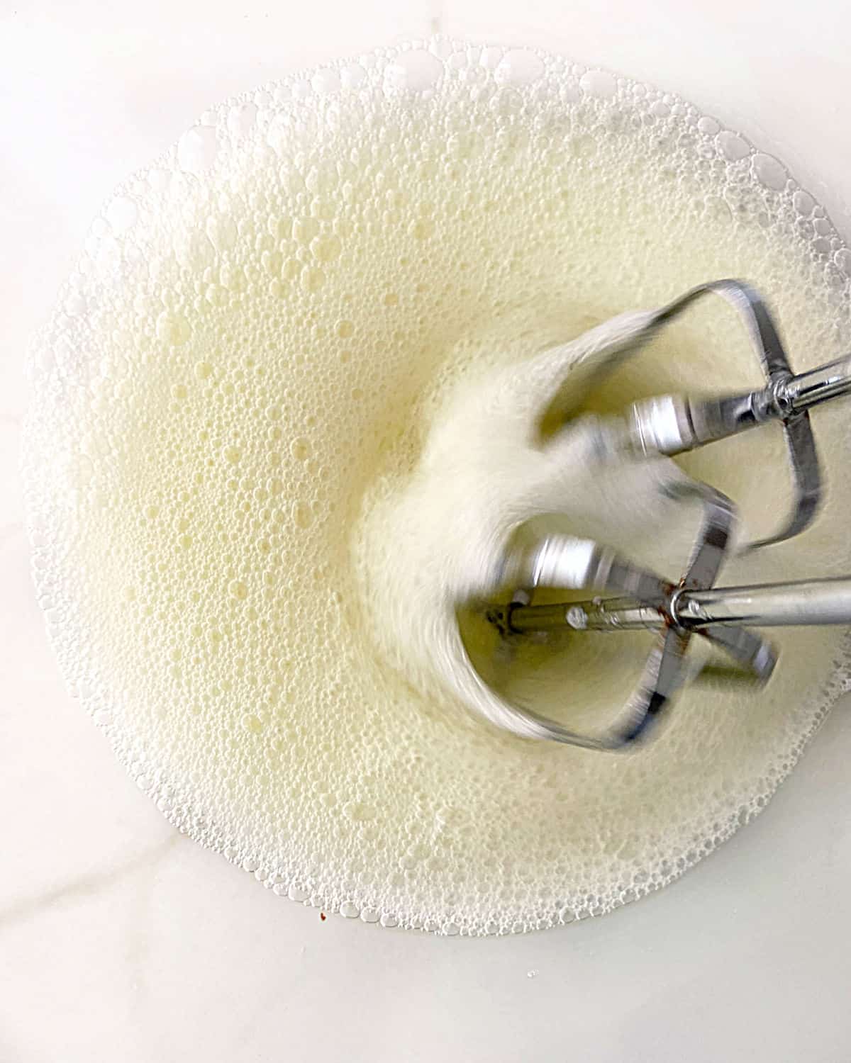 Frothy egg white mixture being beaten in a glass bowl on a white marble surface.