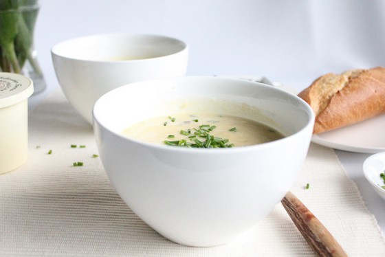 White bowl with leek and potato soup with chives, another bowl and plate with bread in background. 