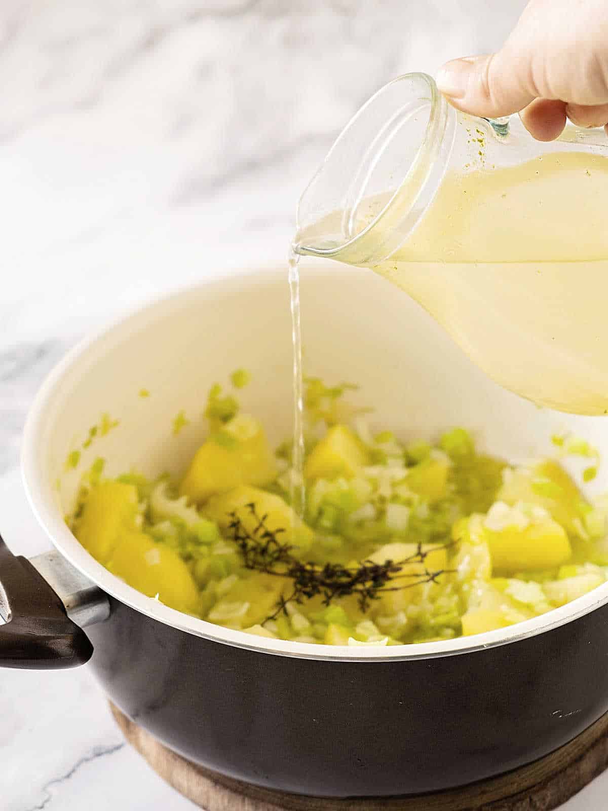 Adding broth from a jug to a pot with potatoes and leeks.