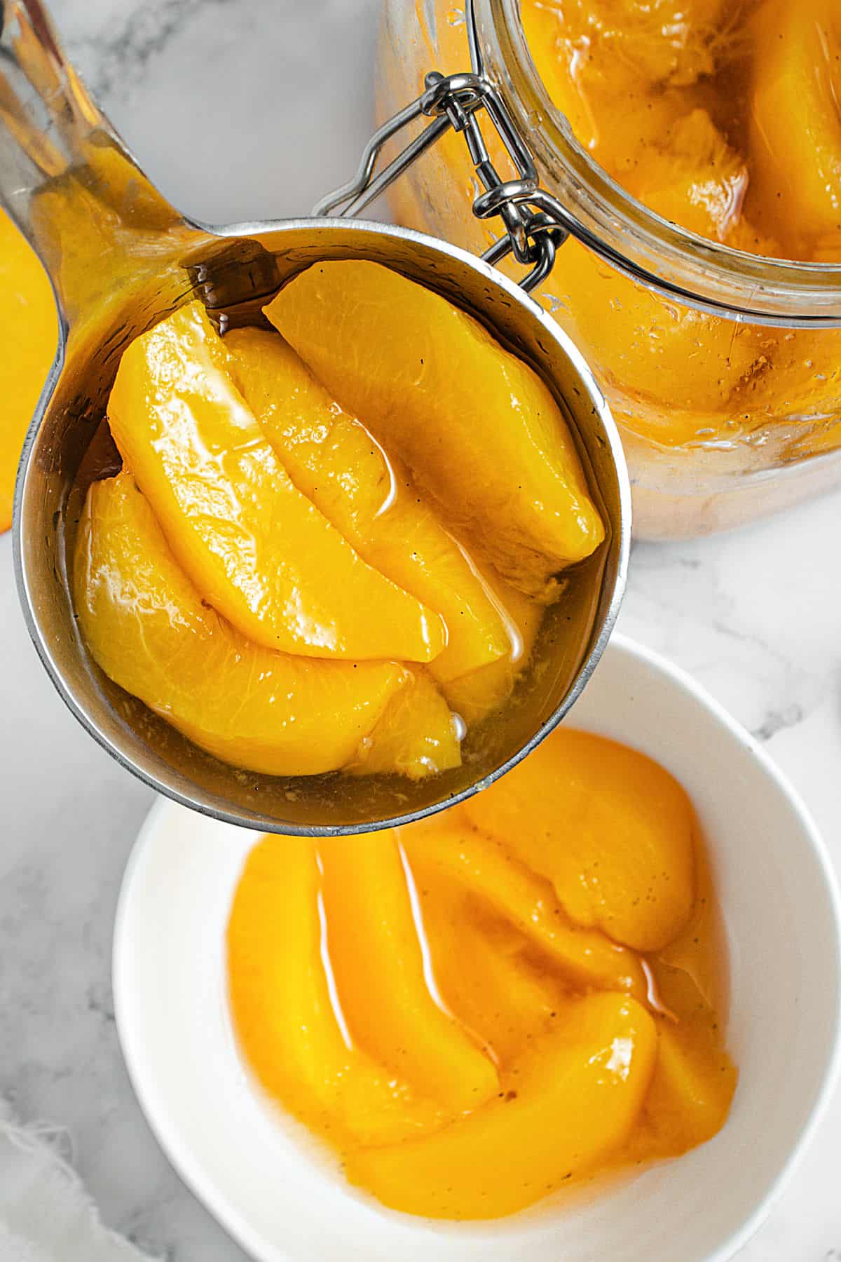 A metal ladle with peach slices in syrup. White bowl and jar beneath. Marble surface.