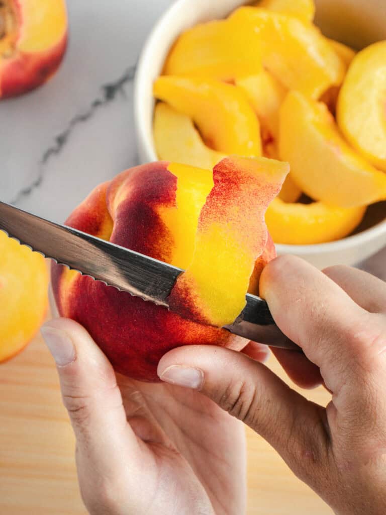 Peeling a fresh peach over a white bowl with slices.