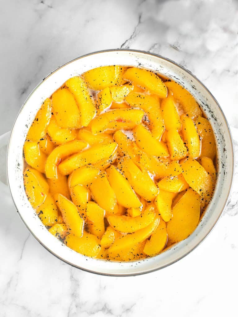 Top view of white saucepan with cooked peaches in syrup. Gray marbled surface.