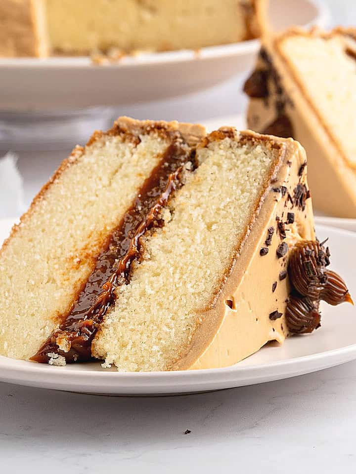 Slice of vanilla dulce de leche cake on a white plate. More cake in the background.