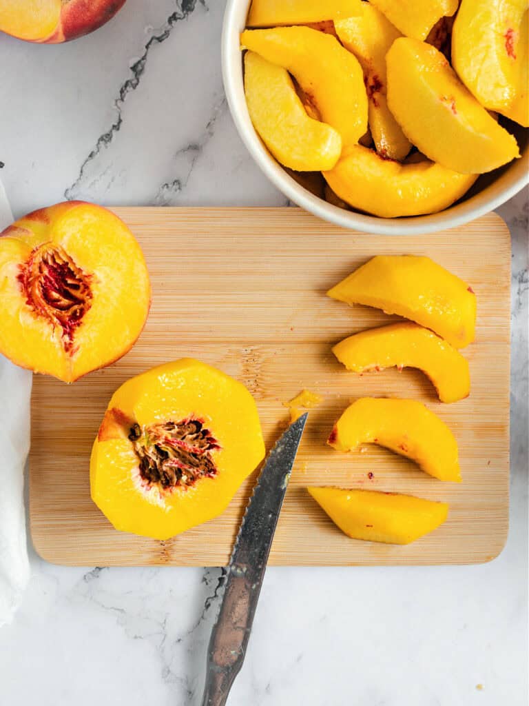 Wooden board on marbled surface with sliced and halved peaches. White bowl with more pieces.