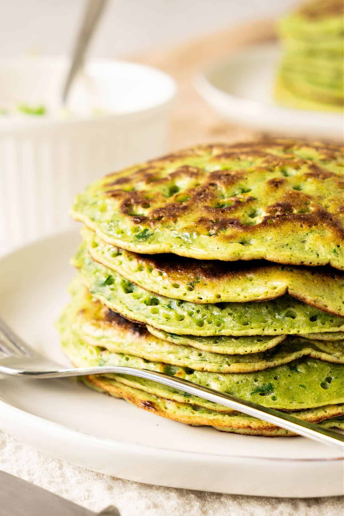 Stack of chard pancakes on a white plate. Silver fork. White bowl in the background.