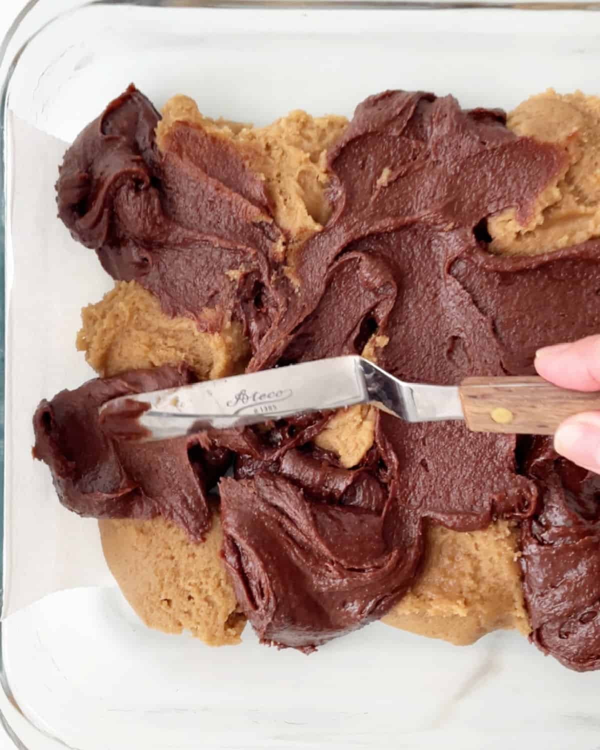 Vanilla and chocolate cookie dough being marbled with an offset spatula in a glass baking dish.