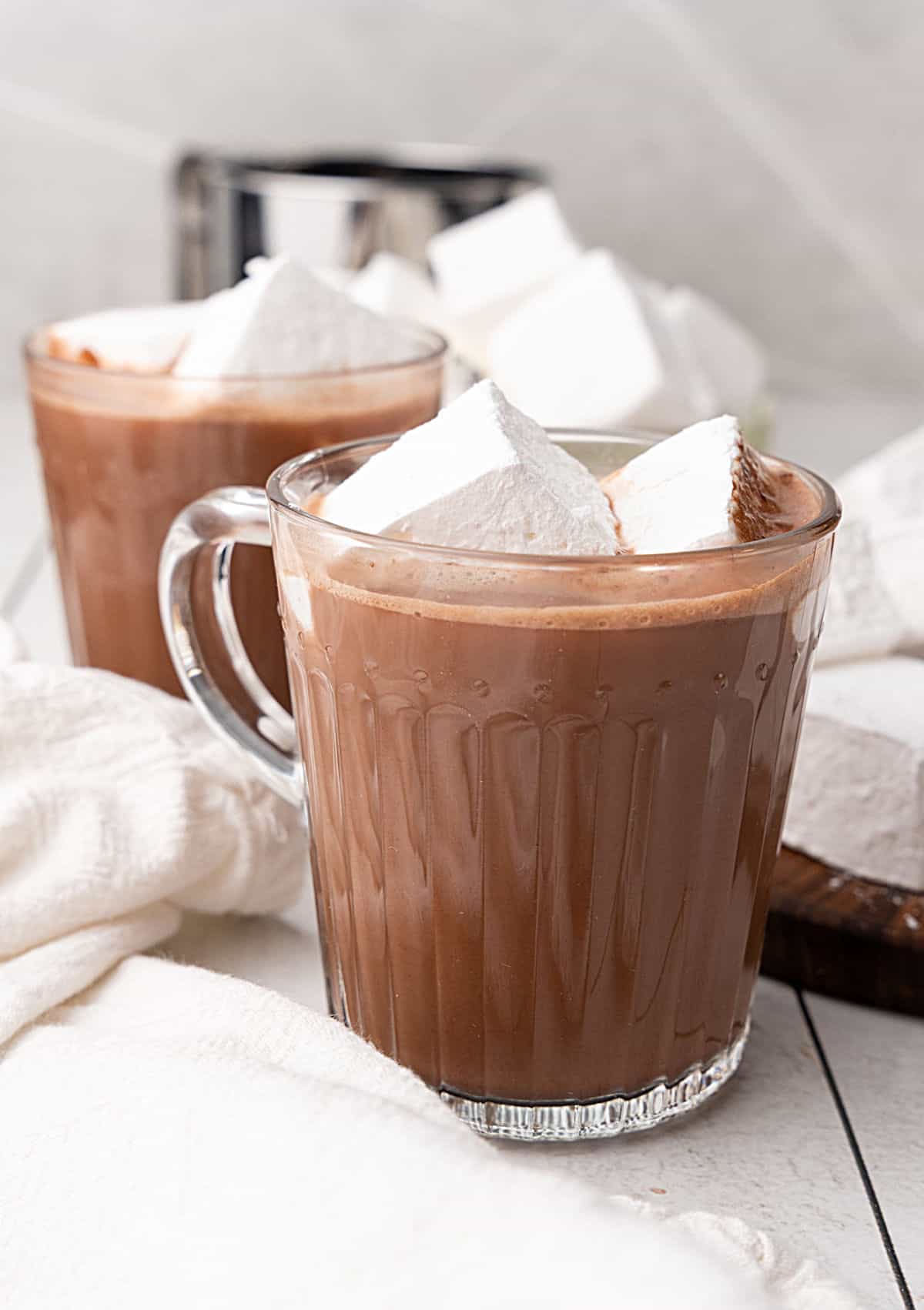 Two hot chocolate glass mugs with marshmallows. White cloth and surface, grey background.