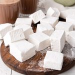 Vanilla marshmallows piled on a wooden board with hot chocolate mugs in the background.