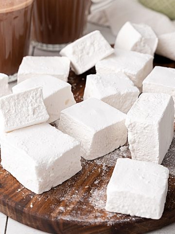 Vanilla marshmallows piled on a wooden board with hot chocolate mugs in the background.