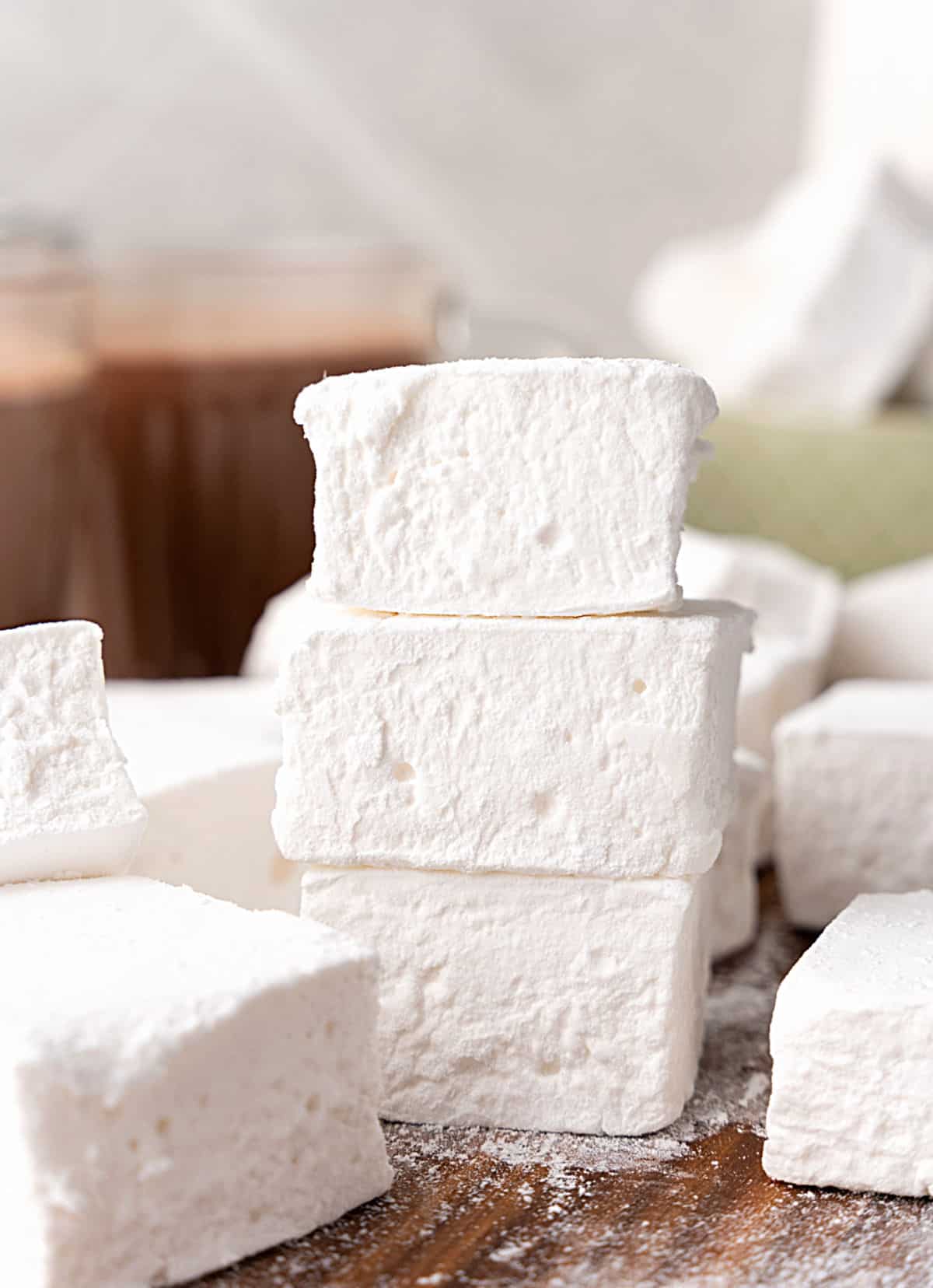 Stack of marshmallows amid many others on a wooden board. Hot chocolate in the grey background.