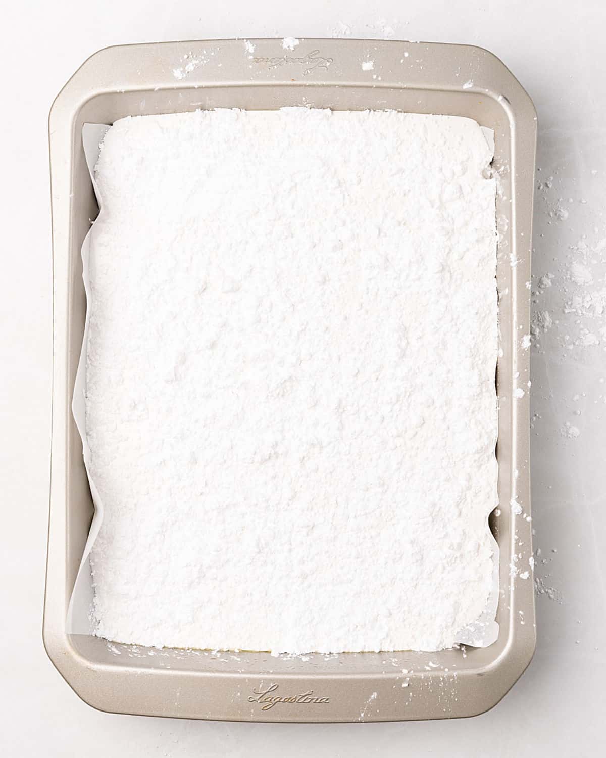 Powdered sugar coated marshmallow mixture in a parchment-lined baking pan on a white surface.