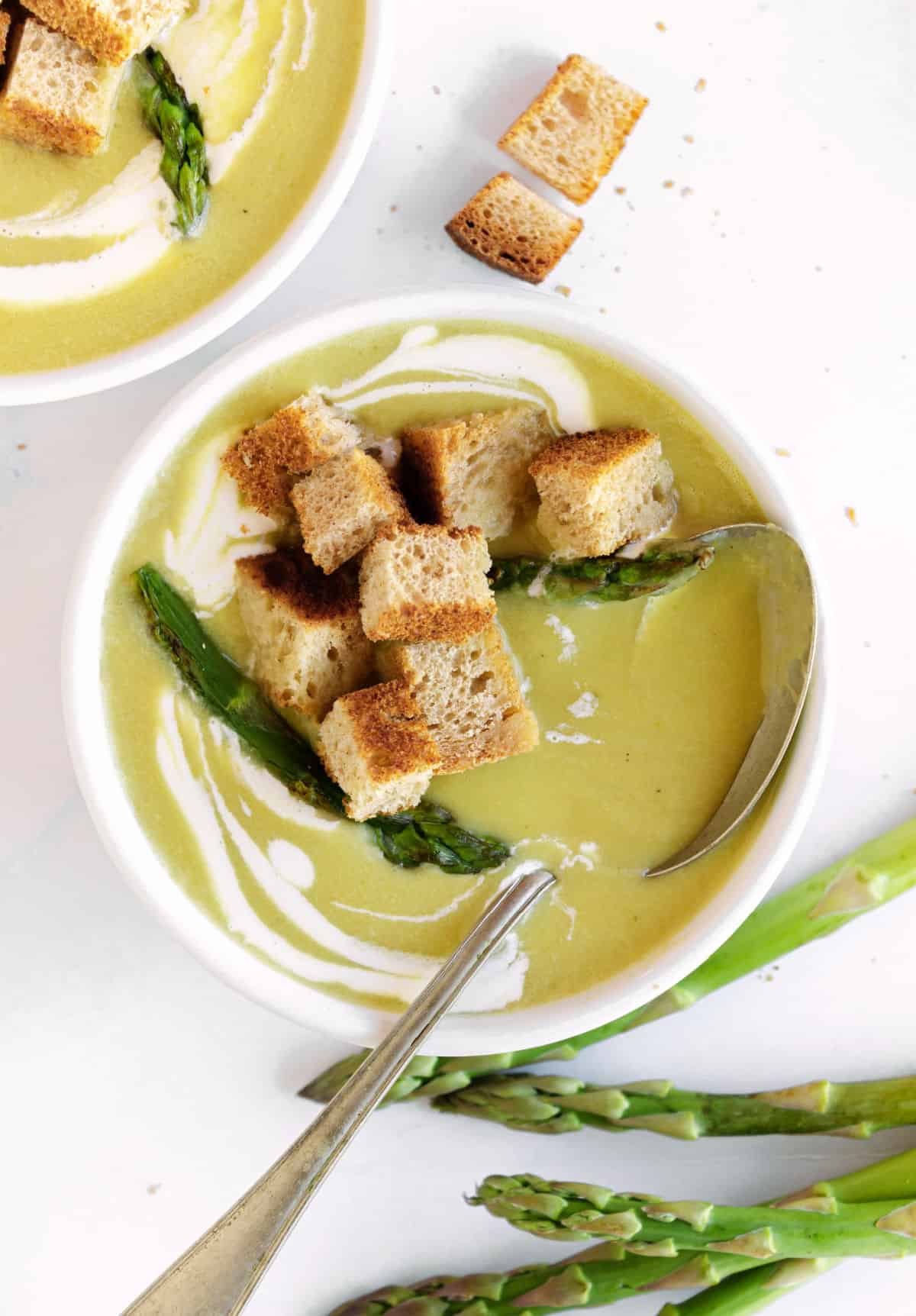 Croutons-topped white bowl of asparagus soup. Silver spoon. White surface.