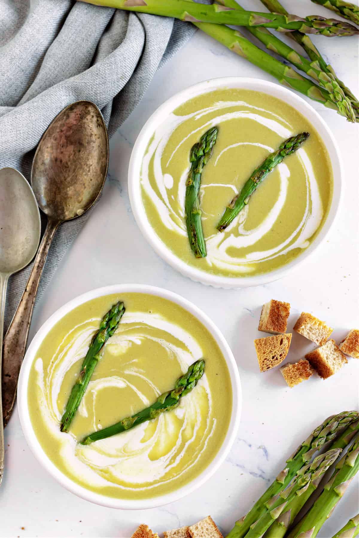 Two white bowls with asparagus soup. White surface, croutons, spoons, fresh asparagus.