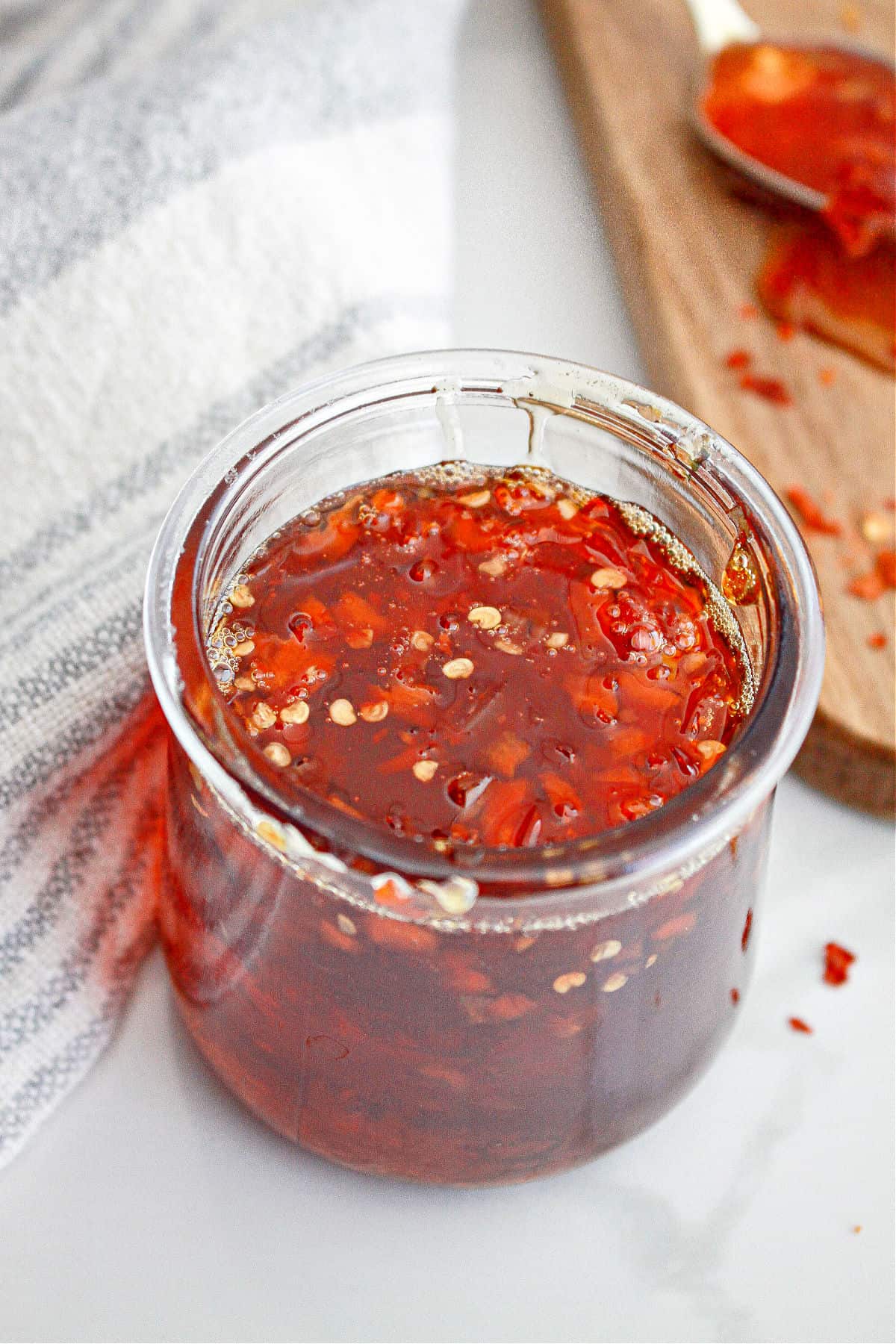 White surface with red chili jam in a jar. Striped cloth, wooden board. 