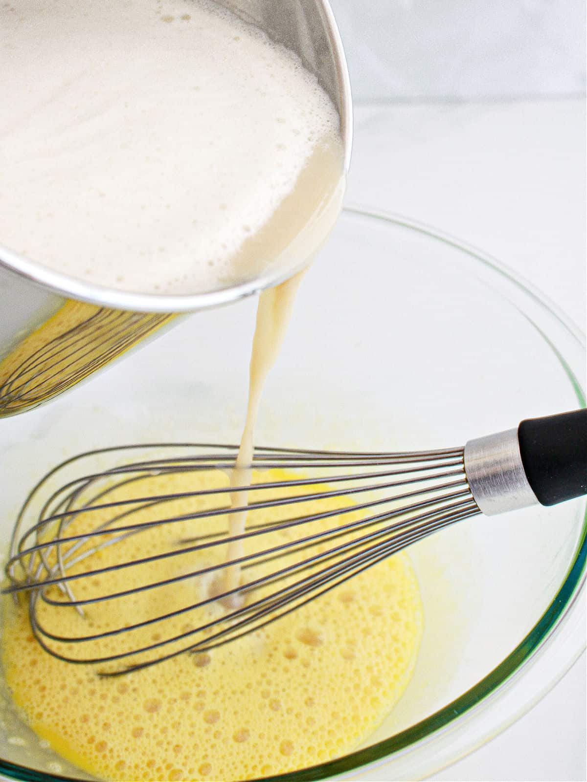 Pouring milk into beaten egg yolks in a glass bowl, a whisk. White background.