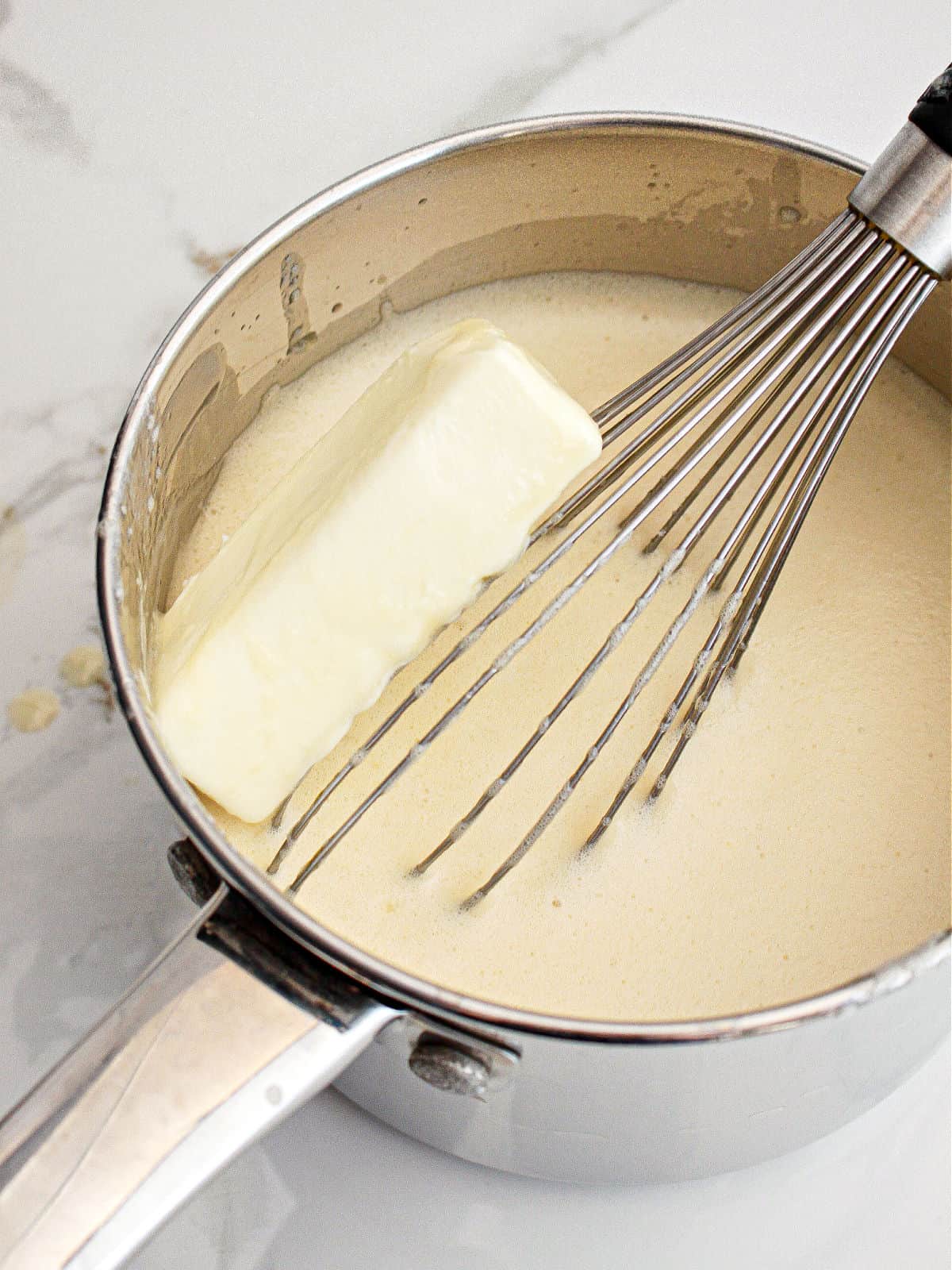 Butter added to a metal saucepan with a custard mixture and a whisk. White background.