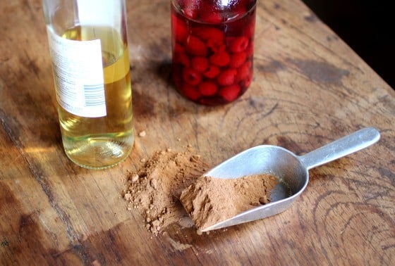 Wooden table with bottle of white wine, macerating raspberries, and metal scoop with cocoa powder.