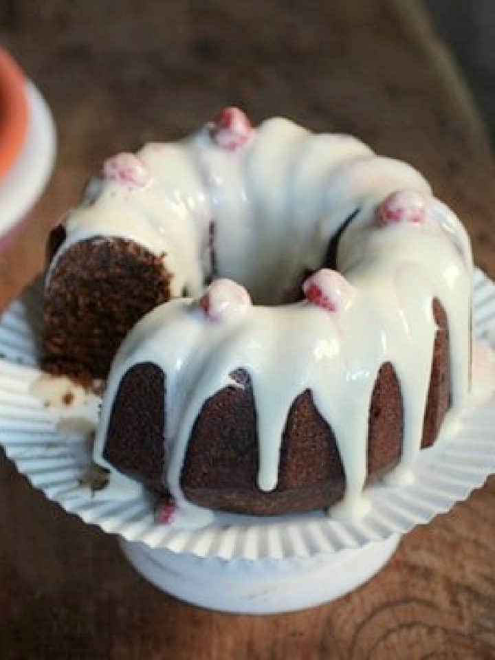 White glaze over chocolate bundt cake on a white cake stand on a wooden table.